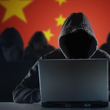 A hooded figure works on a laptop in front of a Chinese flag, symbolizing the online scammers behind the fake crypto investment scheme that defrauded a 60-year-old Hong Kong woman of $2.7M–$2.8M. The image visually represents the anonymous cybercriminals who lured the victim into sending additional deposits to "unlock" withdrawals on a fraudulent platform.