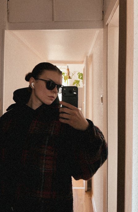 Mirror selfie of a person wearing dark sunglasses, earbuds, and a dark plaid hooded jacket, holding a smartphone in a hallway with a plant visible in the background.