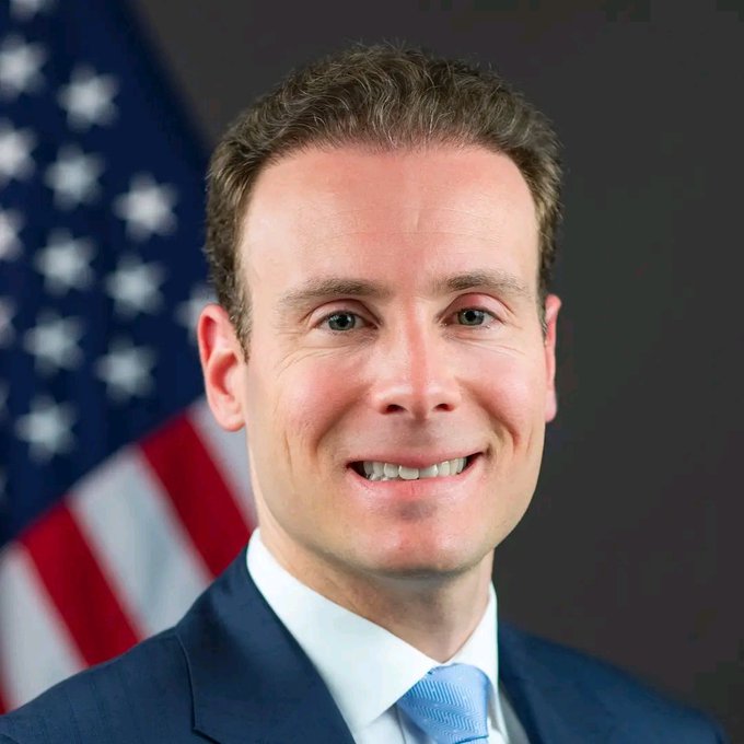 A man in a navy suit and light blue tie smiles in front of an American flag, consistent with an official portrait of CFTC Chair Mike Selig, who is featured in the post discussing the CLARITY Act's progress for crypto regulation.
