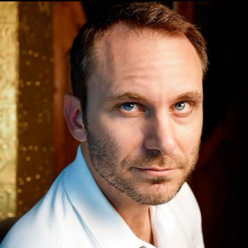 A close-up portrait of a man with short brown hair, light stubble, and striking blue eyes, wearing a white shirt, photographed against a warm-toned indoor background.
