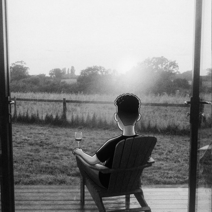 A black and white photo showing a person from behind, sitting in an Adirondack chair on a deck holding a wine glass, looking out at a rural field at sunset.