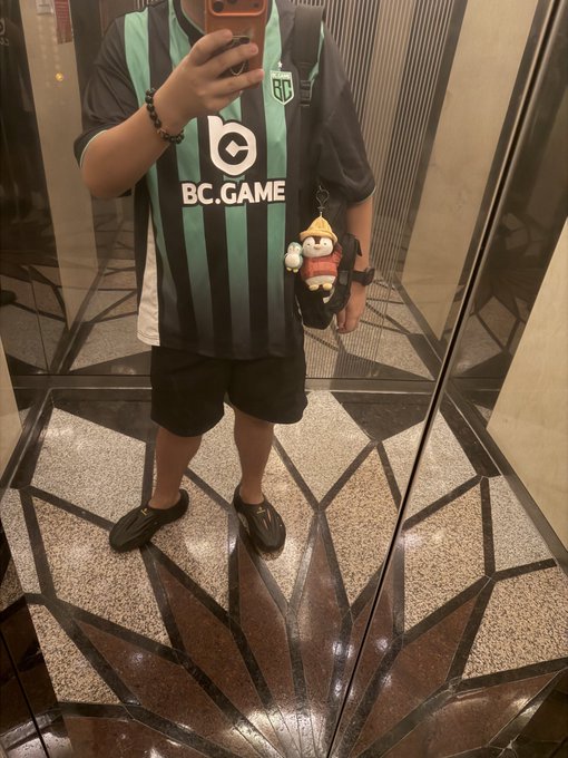 Mirror selfie of a person wearing a green and black striped BC.GAME soccer jersey, black shorts, and a small plush keychain charm.