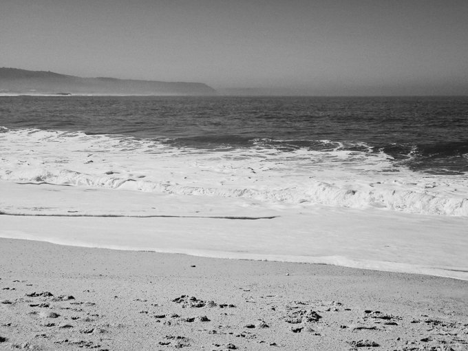 A black-and-white photograph showing gentle waves washing onto a sandy beach, with a faint coastline visible on the horizon under a clear sky.