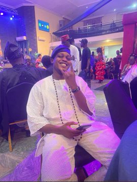 A man dressed in traditional white Nigerian attire and a blue cap sits in a chair at an indoor event, smiling and gesturing with his hand.