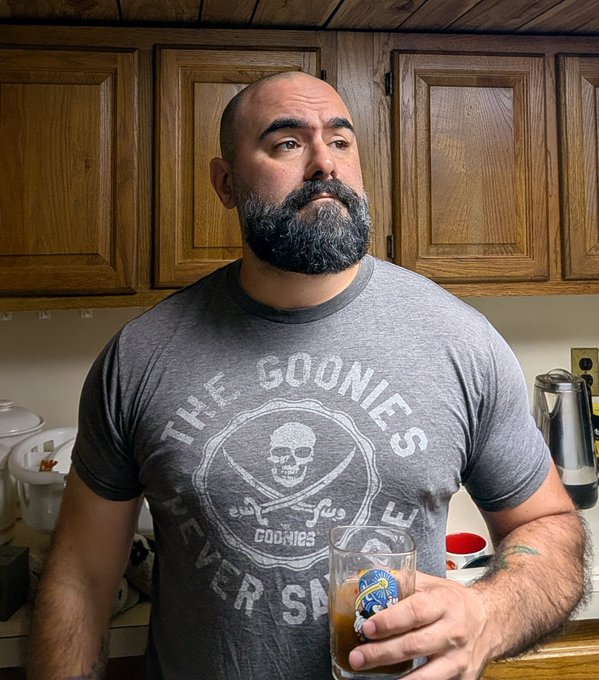 A bald man with a full dark beard wearing a grey 'THE GOONIES NEVER SAY DIE' skull t-shirt, holding a glass of beer with a 'Helgas' logo while standing in a kitchen.