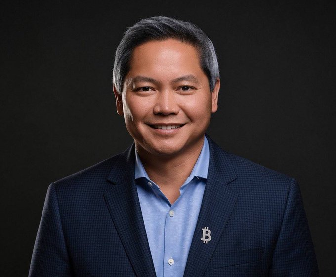 A professional headshot of a smiling man in a dark blazer and blue shirt with a Bitcoin (₿) lapel pin.
