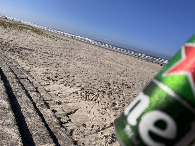 A first-person view on a sandy beach holding a green Heineken beer can in the foreground, with the ocean and shoreline visible in the background.