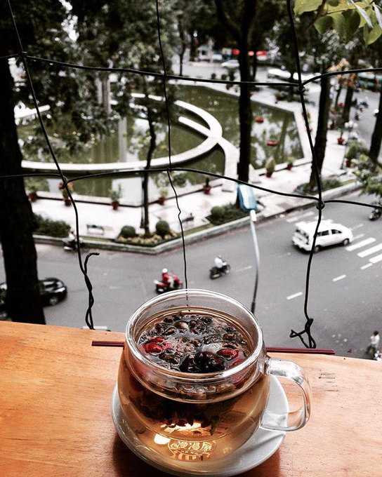 A glass mug of herbal tea with goji berries sits on a wooden ledge overlooking a city street with a scenic pond and park below, framed by hanging string lights—perfectly capturing the "chill, coffee and view" Sunday vibe from the post.