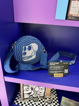A blue circular crossbody bag featuring a cartoon character design displayed on a purple shelf, with a tag reading 'MOKU MURA LIFE' and a price of ¥4,950.