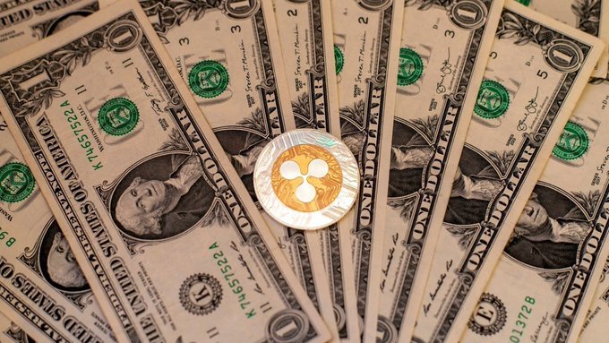 A fan of fanned-out one-dollar bills featuring George Washington's portrait, with a physical Ripple (XRP) gold coin resting in the center—visually reinforcing the bold analyst prediction of XRP reaching $499.98 within three months.