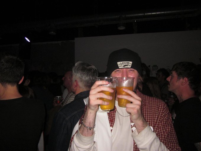A photo of a young man wearing a Vans beanie and a checkered shirt, holding up two plastic cups of beer in a dark, crowded setting.
