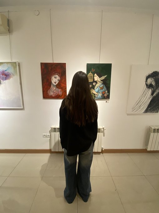 A woman with long dark hair, seen from behind, stands in an art gallery viewing several paintings hung on a white wall above a radiator.