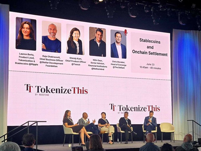 The image shows a group of people sitting around a table with a large screen behind them displaying a presentation about tokenization. The panel consists of six individuals seated at the table, engaged in discussion or listening to the presentation. The text on the screen reads "Wherever you see embedded friction when money is moving from point A to B, we see value." This suggests that the group might be discussing how blockchain technology can create value by reducing friction and streamlining processes related to financial transactions.