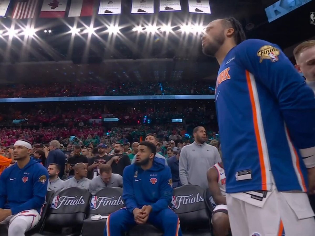 The image shows a basketball player wearing a blue jersey with orange stripes, sitting down in a stadium full of people watching a game. There are several other players visible in the background as well. The player's face can be seen looking up at something or someone outside the frame. The surrounding crowd appears to be engaged and focused on the game, creating an atmosphere of excitement and anticipation for the upcoming play.