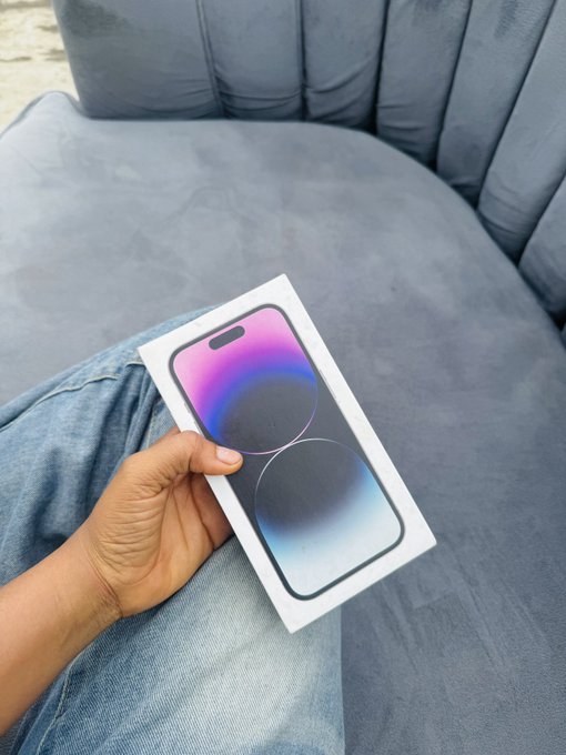 A hand proudly displays a new iPhone box over denim-clad legs on a gray couch, likely the iPhone 15 Pro (visible on the box artwork), marking the addition of new equipment for Crypto Street production thanks to Web3.