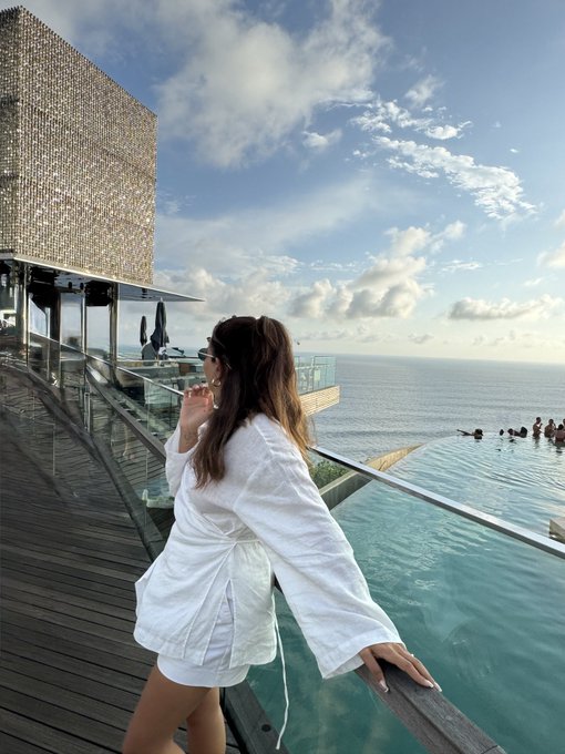 A woman in a white bathrobe leans against a glass railing at a stunning oceanfront infinity pool, gazing out at the sea under a bright, partly cloudy sky. The luxurious resort setting perfectly captures the relaxed weekend vibes of "GM frens Happy Weekend."