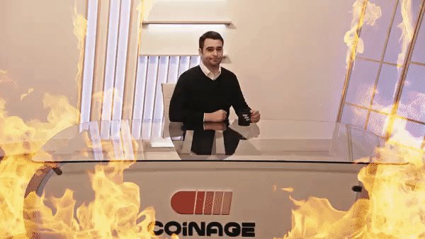 The image shows a man sitting at a desk with a computer, surrounded by flames coming out of an open window behind him. The fire appears to be burning something or someone inside the room. The background features a white wall that contrasts with the flames, creating a dramatic and intense scene. The post includes text about cryptocurrency crashes and mentions Bitcoin, which is likely related to the current situation in the image.