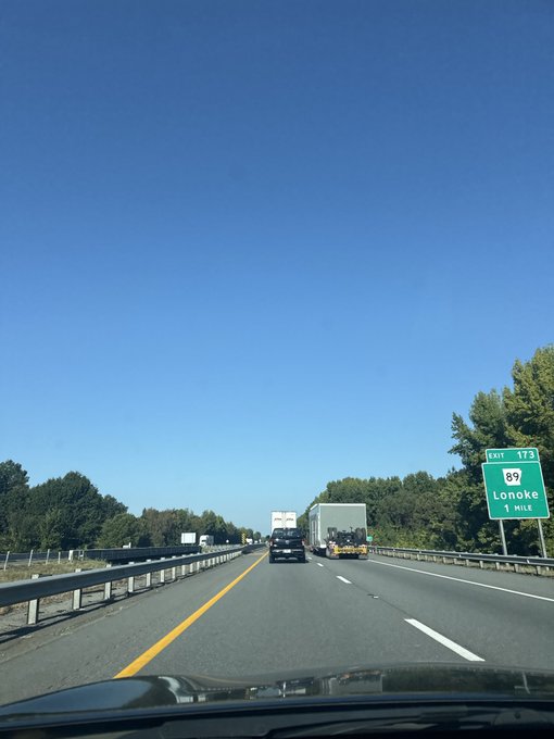 A view from inside a car driving on a highway under a clear blue sky, with a green highway sign indicating 'EXIT 173' for Route 89 to Lonoke in 1 mile.