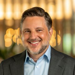 A smiling middle-aged man with a beard, wearing a dark suit jacket and light blue shirt, poses in front of a blurred golden bokeh background. The portrait aligns with the post featuring Grayscale CEO Peter Mintzberg discussing bullish signs for crypto's recovery.