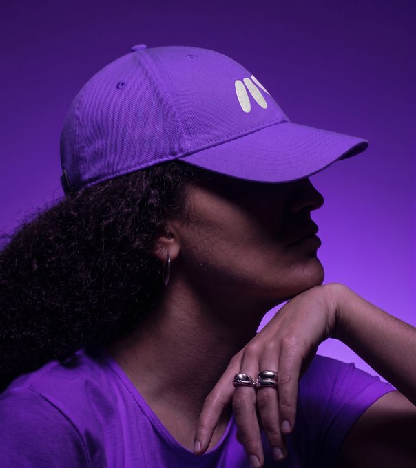 The image showcases a stylish side profile of a person wearing a vibrant purple baseball cap featuring two white slash marks as a logo, paired with a matching purple t-shirt. Their curly hair frames their face, and they sport silver hoop earrings and multiple silver rings on their fingers, with one hand resting thoughtfully near their chin. The dramatic purple lighting and gradient background create a bold, monochromatic aesthetic that perfectly aligns with the promotional announcement for an Africa Block Festival giveaway in Nairobi, Kenya, offering a ticket and merchandise prize to be awarded at 8pm EAT, with the model's fashionable, branded look hinting at the event's trendy, streetwear-inspired merchandise available to winners.