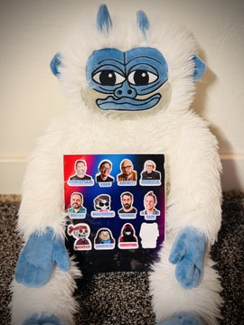 A white and blue fluffy plush toy with a blue face holds a card featuring stickers of various soccer players.