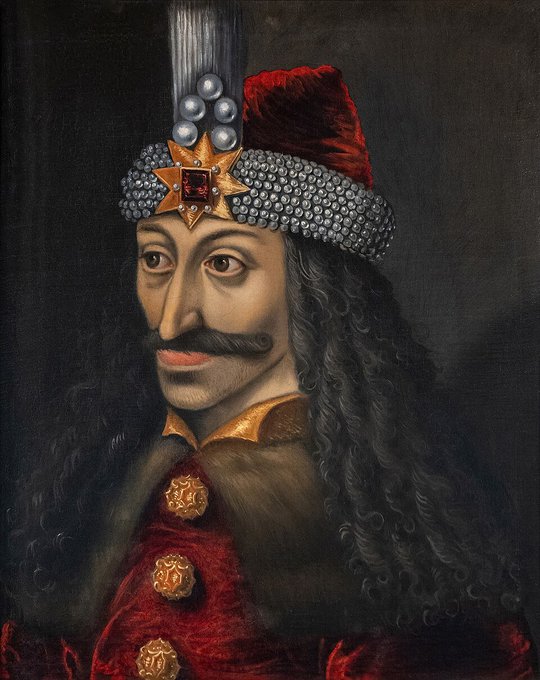 The image features a portrait of a man with long, curly hair wearing a red coat and a gold hat adorned with a large gemstone. He has a mustache and a beard, giving him a distinguished appearance. The background appears to be black, which contrasts with the vibrant colors of his attire. The caption "Bring back Vlad" suggests that this image might be related to a historical event or figure named Vlad, possibly from the Roman era.
