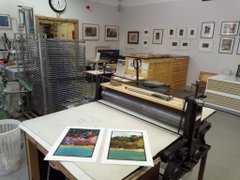 Two prints sit on a worktable in a printmaking studio, one featuring an autumn scene with vibrant orange foliage and the other depicting a watery landscape, awaiting their unveiling after arrival.