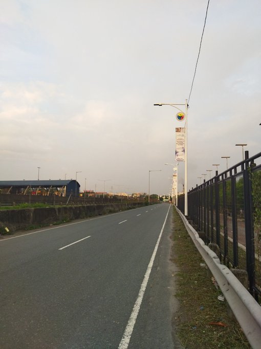 An empty road stretches into the distance with streetlights along the side, one displaying a banner, under an overcast sky.