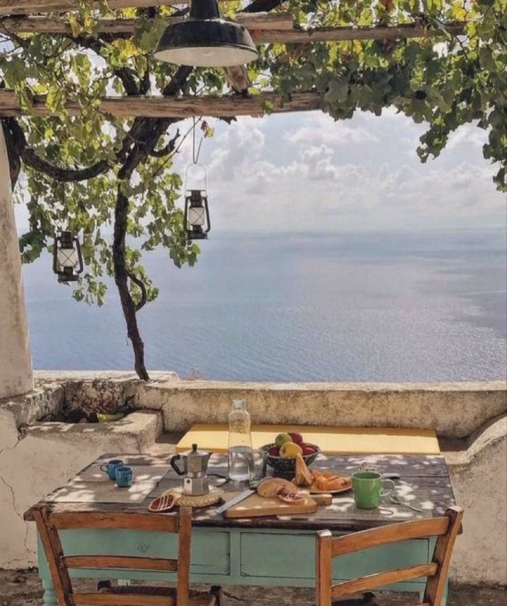 A rustic outdoor table set with coffee, croissants, and fresh fruit overlooks a serene Mediterranean seascape beneath a leafy grapevine pergola, embodying the post's quiet message that true richness is found in peaceful, simple moments rather than luxury.