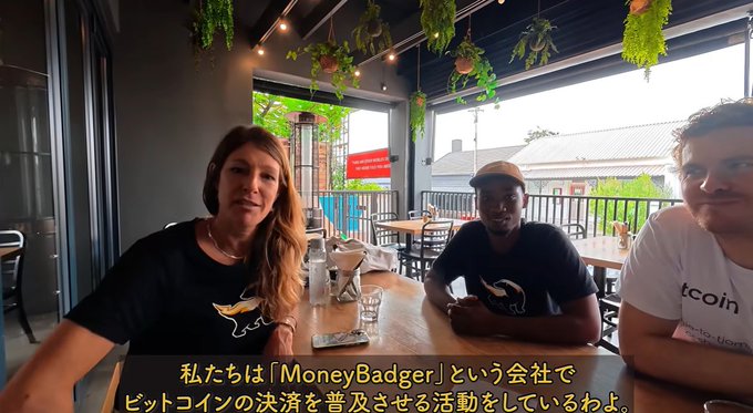 Three people sitting at a table in a cafe-like setting with Japanese subtitles translating to 'We are working to spread Bitcoin payments with a company called MoneyBadger.'