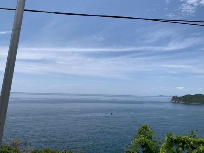 A serene ocean view with calm blue water stretching to the horizon under a partly cloudy sky, with a utility pole visible on the left and green foliage in the foreground.