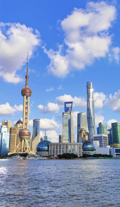 A scenic view of the Shanghai skyline featuring the Oriental Pearl Tower and other skyscrapers along the waterfront under a partly cloudy blue sky.