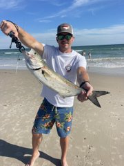 A man wearing a white t-shirt, colorful board shorts, sunglasses, and a cap stands on a sandy beach holding up a large caught fish.