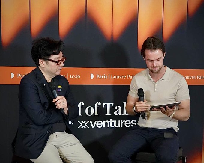 In the image, two men are sitting next to each other at a table, both holding microphones as they engage in conversation. They appear to be discussing something related to the "OF TAL" event that took place in Paris in 2016. One of them is wearing glasses and has short hair, while the other man has long hair. The background features an orange banner with white text, which reads "Paris (Lourve Palais) 2016".