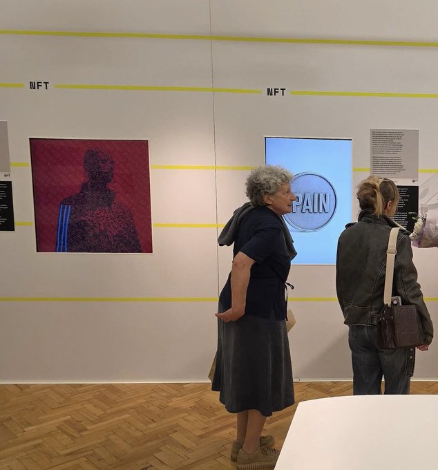 Two women view an NFT exhibition featuring a glitch-style portrait on the left and an illuminated screen displaying the word 'PAIN' on the right.
