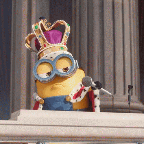 A Minion character dressed as a king, wearing an ornate gold and pink crown and a royal robe, standing at a podium with a microphone in front of classical columns.