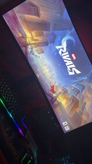 A computer monitor displaying the Marvel Rivals loading screen, with an RGB keyboard visible on a desk in a dimly lit room.
