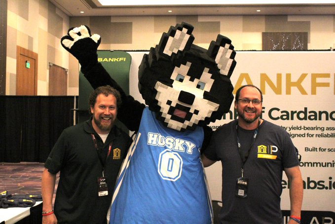 Two smiling men pose on either side of a large Husky mascot wearing a blue jersey; a 'BANKFI' booth is visible in the background.