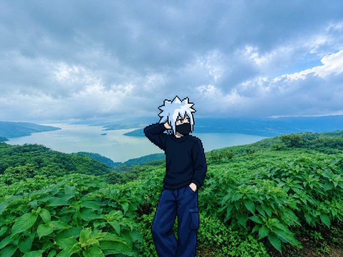 A figure wearing a dark hoodie and dark pants stands on a lush green hillside overlooking a large body of water with distant mountains under a cloudy sky. The person's face has been replaced with an anime-style character mask featuring spiky white hair.