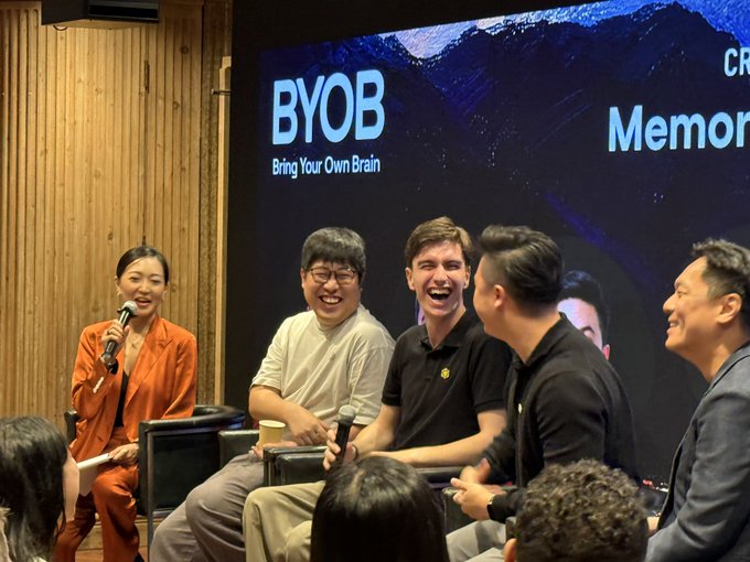 A panel discussion featuring five people seated on stage, with a woman in an orange suit holding a microphone. The screen behind them displays 'BYOB - Bring Your Own Brain' and partial text 'Memory'.