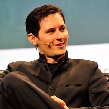 A man with short brown hair wearing a dark jacket and headset microphone smiles while seated, appearing to speak at a panel or interview setting.