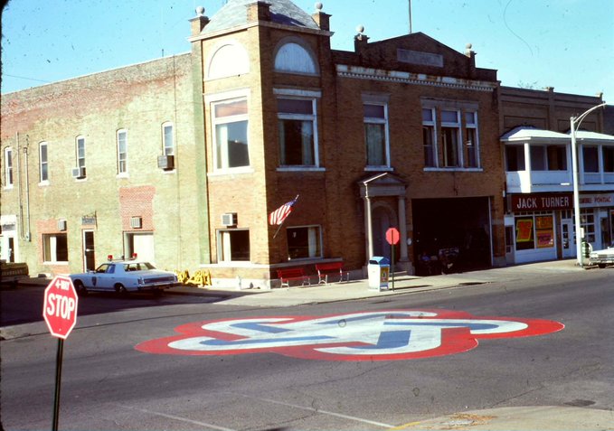 A street scene featuring a fire station with a police car parked outside and a stop sign in the foreground, with a large red, white, and blue 'ZZ' emblem painted on the road.