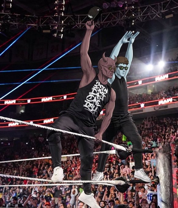 Two cartoon figures wearing devil and alien masks stand on the ropes of a WWE wrestling ring in front of a cheering crowd, with one wearing a shirt reading 'Omega Crew 44'.