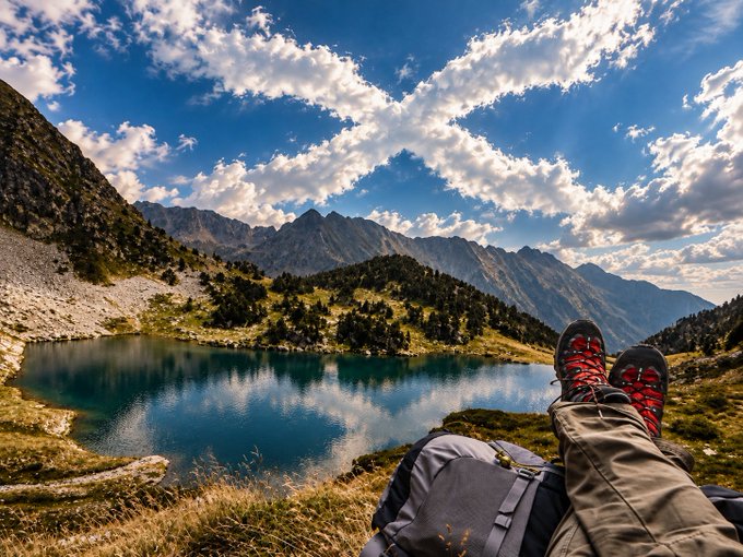 A scenic landscape photograph shows a hiker's legs and hiking boots resting on the grass in the foreground, with a stunning view of an alpine lake and mountain peaks under a partly cloudy sky.