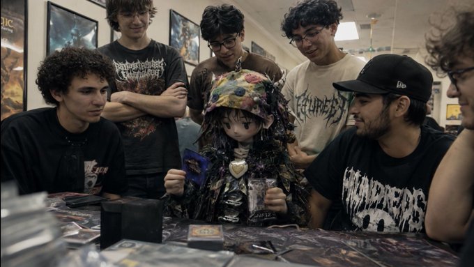 A group of young men in casual clothing and graphic t-shirts gather around a table, looking at cards and items in what appears to be a game or trading card shop.