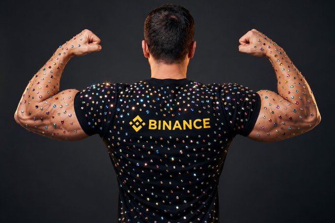 A man facing away flexing his back muscles, wearing a black t-shirt with 'BINANCE' logo on the back.