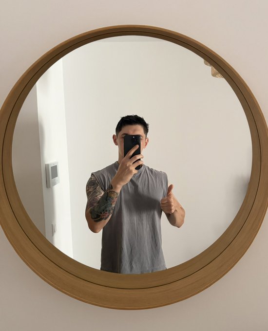A mirror selfie of a young man with a tattooed arm wearing a gray sleeveless shirt, holding a phone and giving a thumbs-up.
