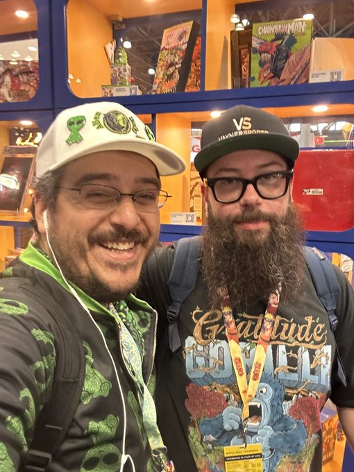 Two men pose together at what appears to be a comic convention; one wears a white cap with green logos and glasses, smiling broadly, while the other sports a long beard, black glasses, and a 'VS' cap along with a 'Gratitude Crew' t-shirt and lanyard.