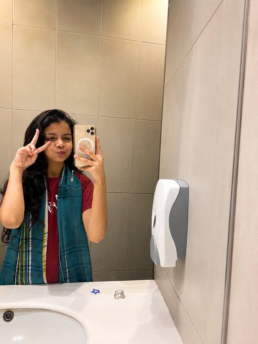 A person takes a mirror selfie in a bathroom, making a peace sign and puckering their lips, wearing a maroon shirt and a blue and green plaid scarf, holding a smartphone with a pop socket.