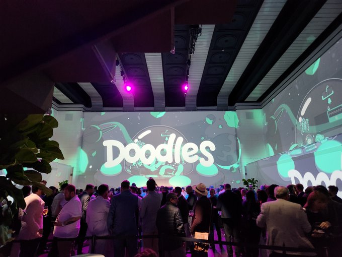 A crowd of people gathered at an indoor event with a large screen projecting the 'Doodles' logo in green.